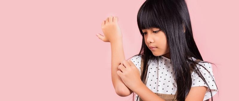 Young girl with dark hair examining her arm against a pink background, wearing a white polka dot top.