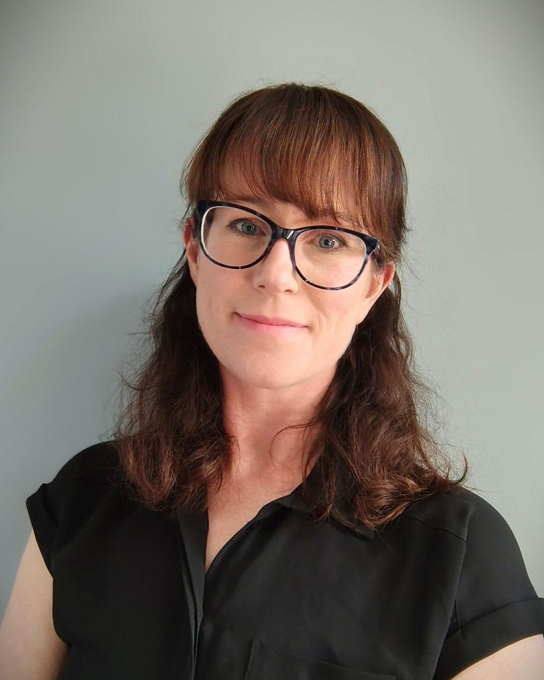Woman with glasses, wavy brown hair and fringe, wearing a black, short-sleeved top.