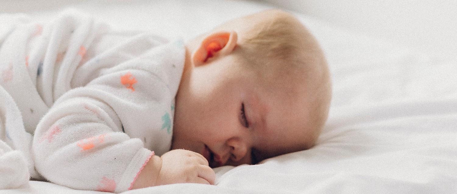 Sleeping infant in white patterned sleepwear resting peacefully on white bedding, with tiny hand curled near face.