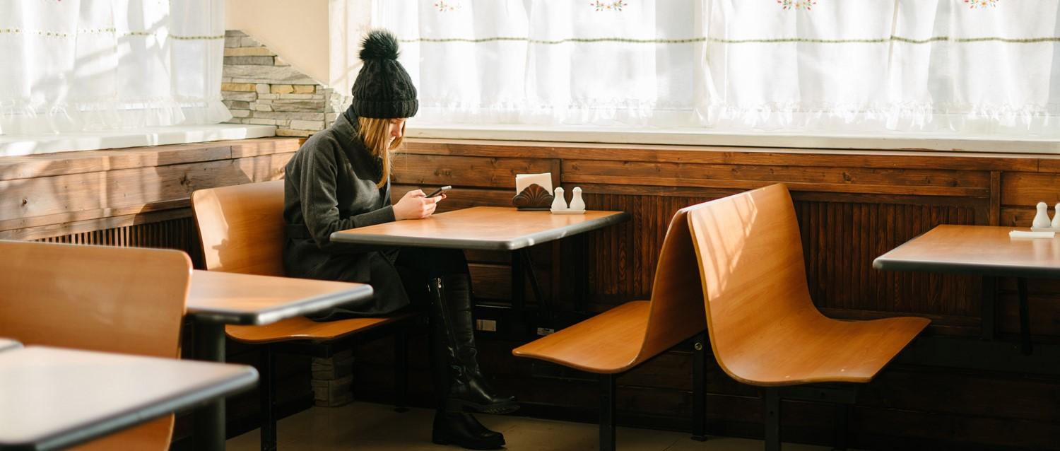 Person in black winter hat and coat using phone at wooden café table with orange chairs by sunlit windows.