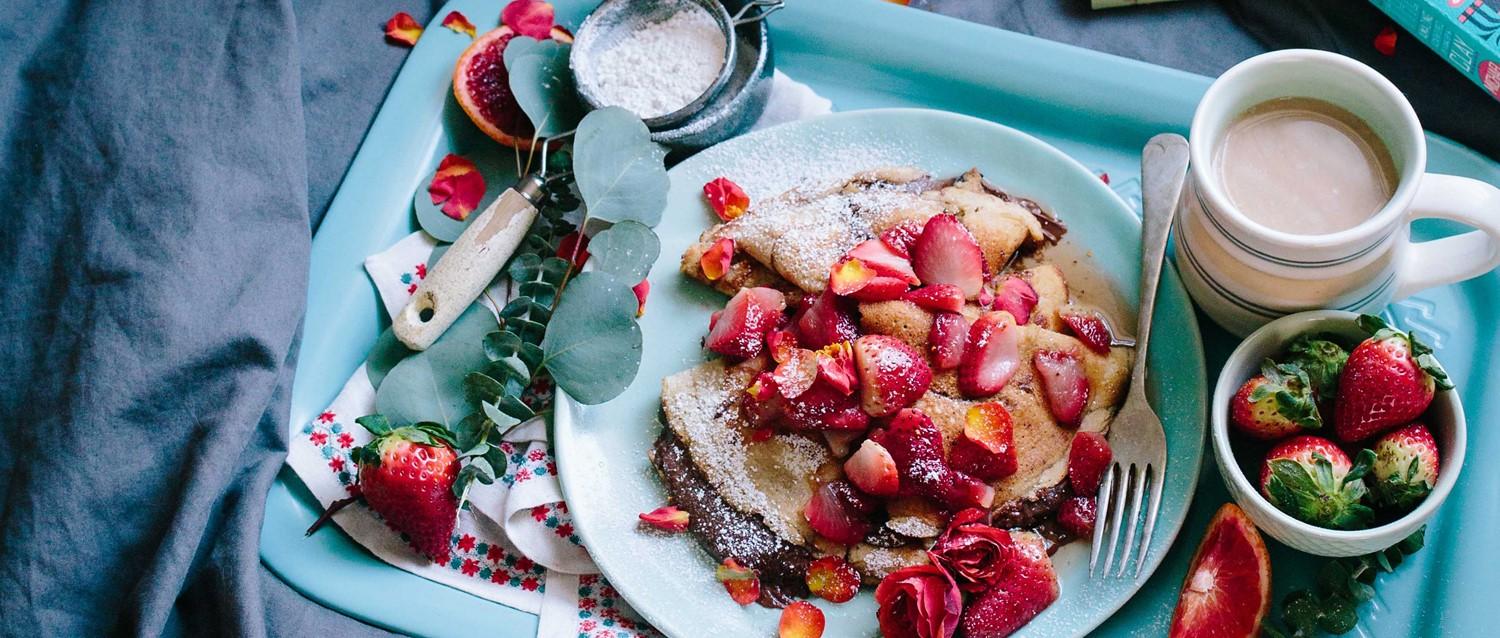 Breakfast tray with crepes topped with fresh strawberries, powdered sugar, coffee, and a small bowl of fruit.