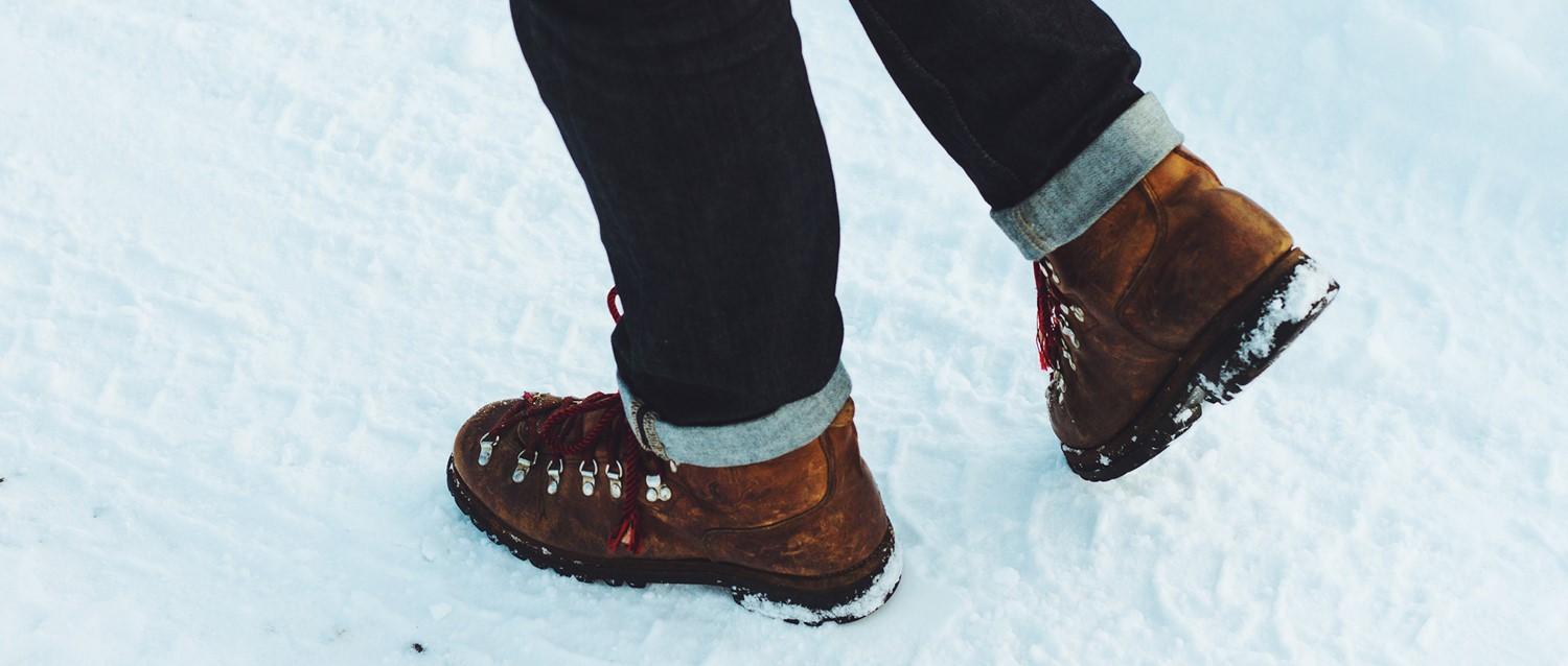 Person wearing brown hiking boots with red laces and rolled-up dark jeans walking through snow.