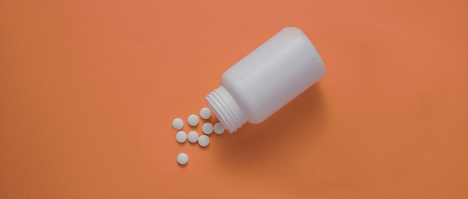 White medicine bottle tipped over with small round pills spilling out onto an orange background.