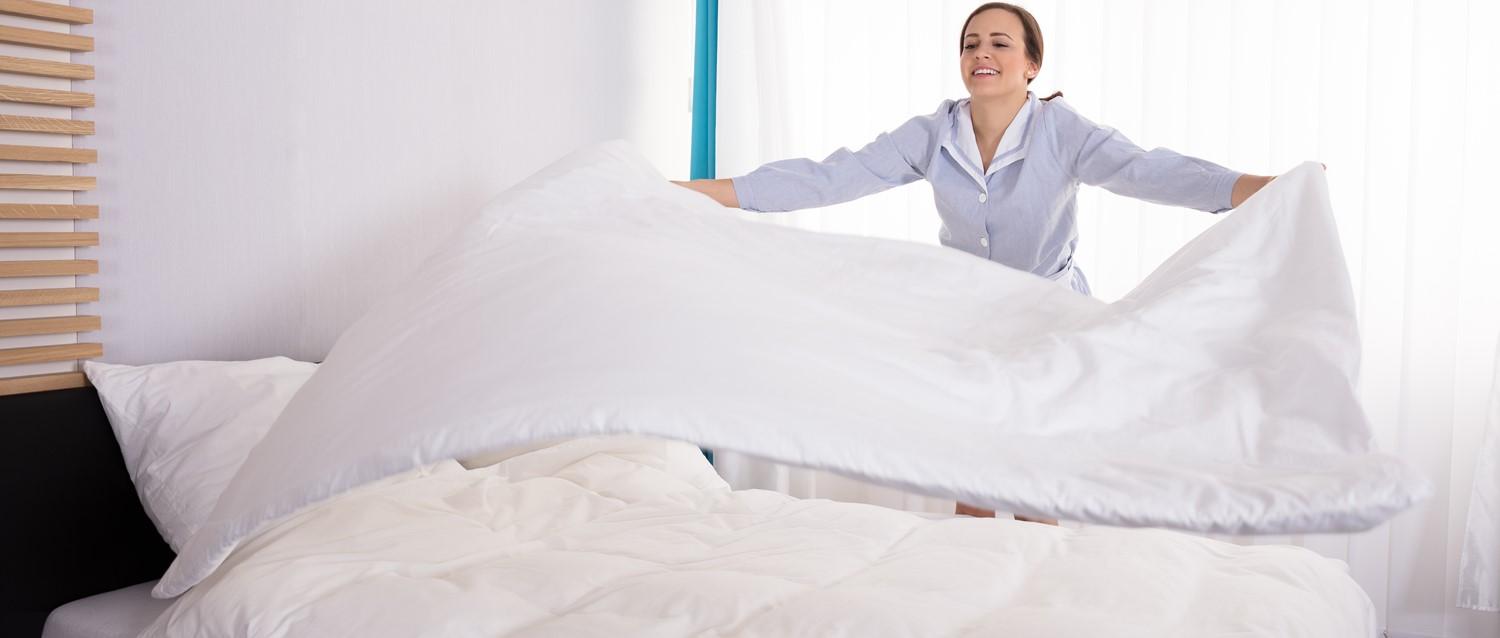 Hotel housekeeper in blue uniform making a bed with white linens in a bright, minimalist room with wooden blinds.