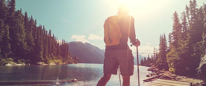 Hiker with walking stick overlooking serene mountain lake at sunset, surrounded by pine forests.